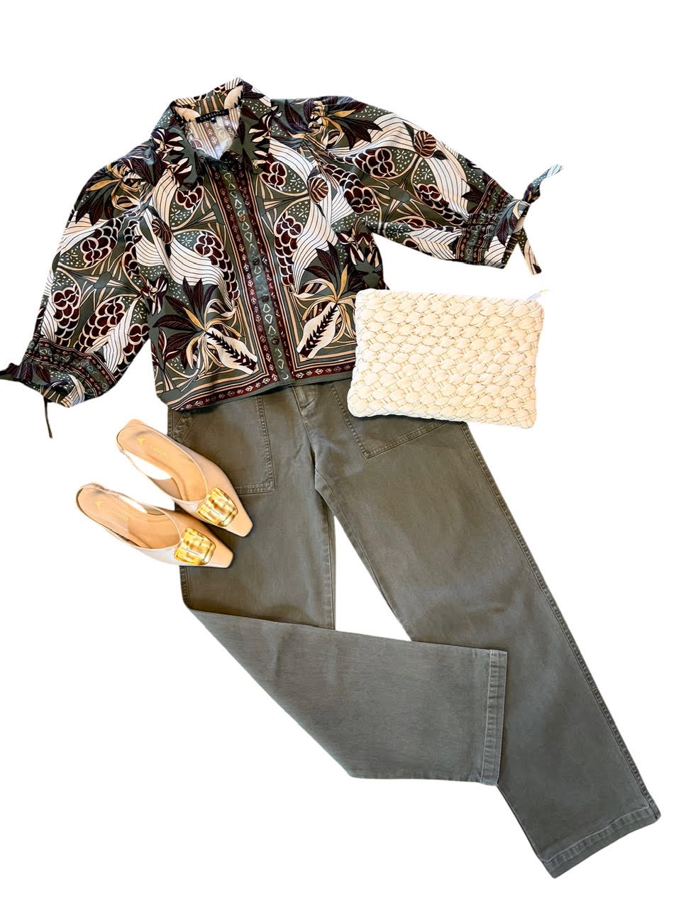 A styled flat-lay outfit featuring a black batik patterned shirt jacket, olive green pants, a cream woven clutch, and beige pointed-toe flats with gold accents, arranged on a solid white background.
