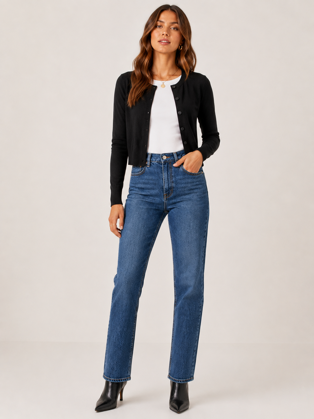 A woman modeling a black cropped cardigan over a white top and blue straight-leg jeans, paired with black ankle boots, against a plain light gray studio background.