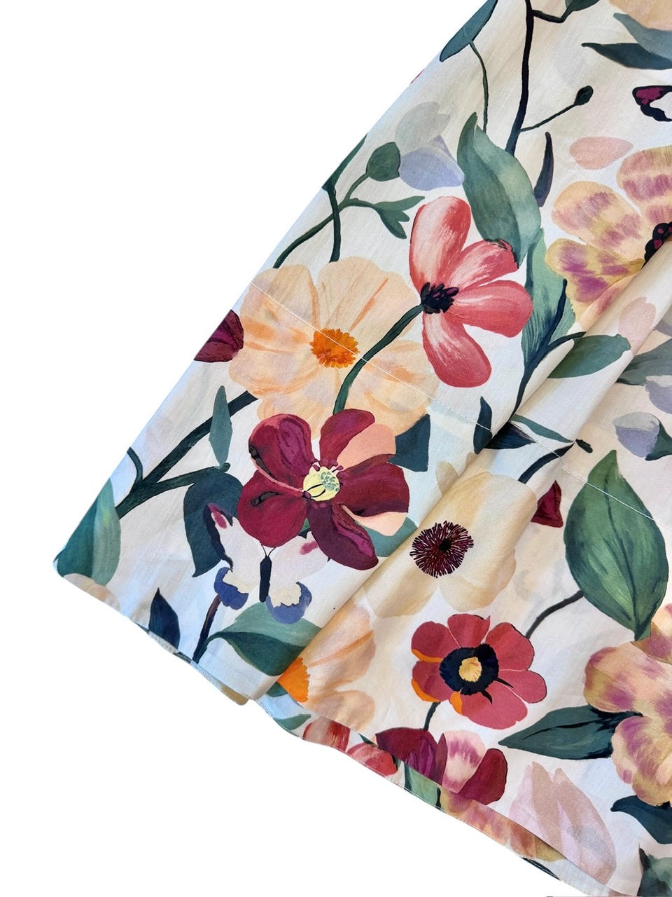 A close-up of a floral fabric with a watercolor-style print of pink, red, peach, and purple flowers with green leaves on a cream background, folded diagonally.