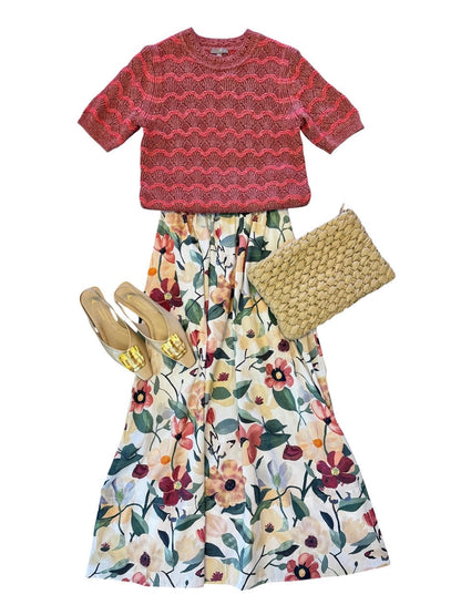 A flat-lay outfit set featuring a short-sleeve red and pink patterned knit top, a long white skirt with a colorful floral print, a woven straw clutch, and gold flat sandals, all arranged on a solid white background.