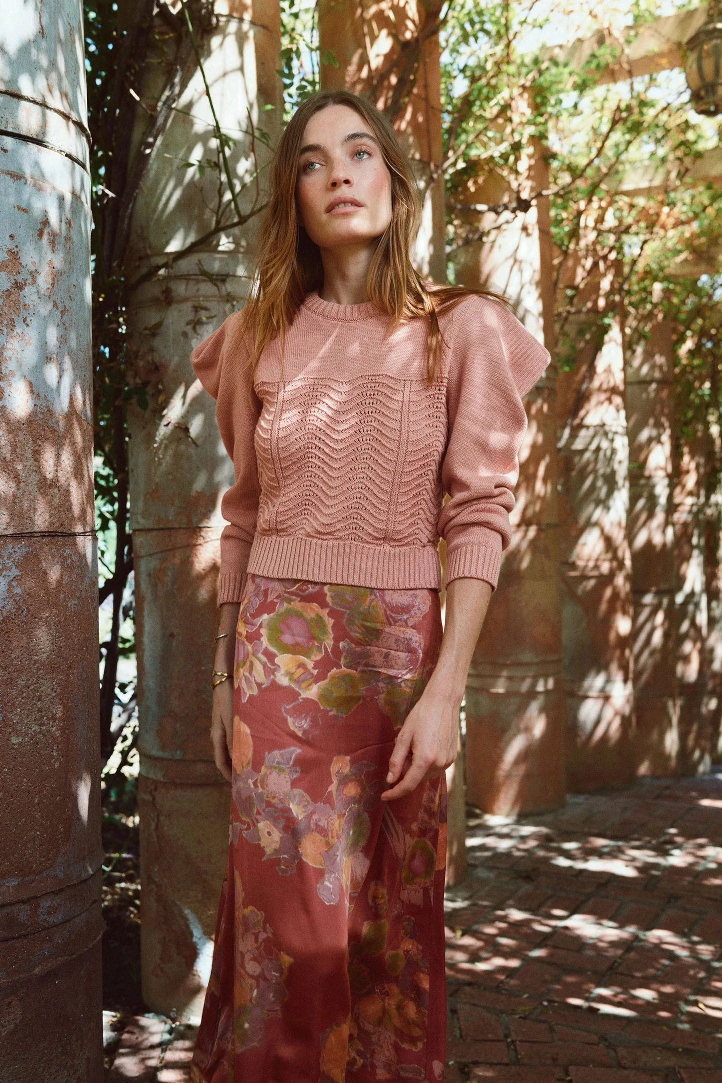 A woman wearing a dusty pink textured knit sweater with puffed shoulders and a rust-red floral maxi skirt, standing in a sun-dappled outdoor corridor with stone pillars.