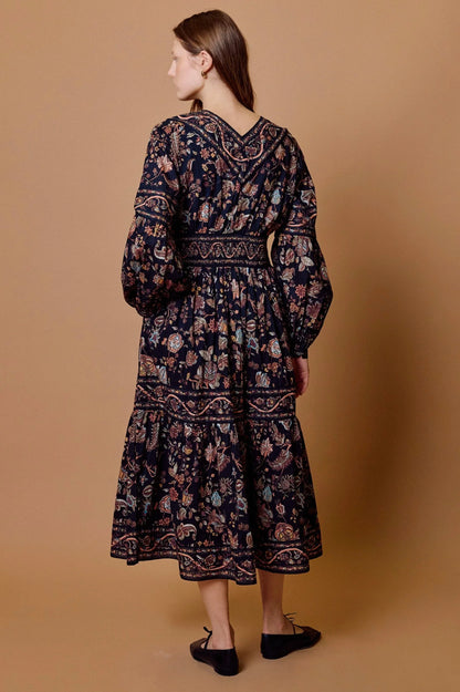 Back view of a woman wearing a printed  longsleeve cotton midi dres paired with brown flats against a tan colored background
