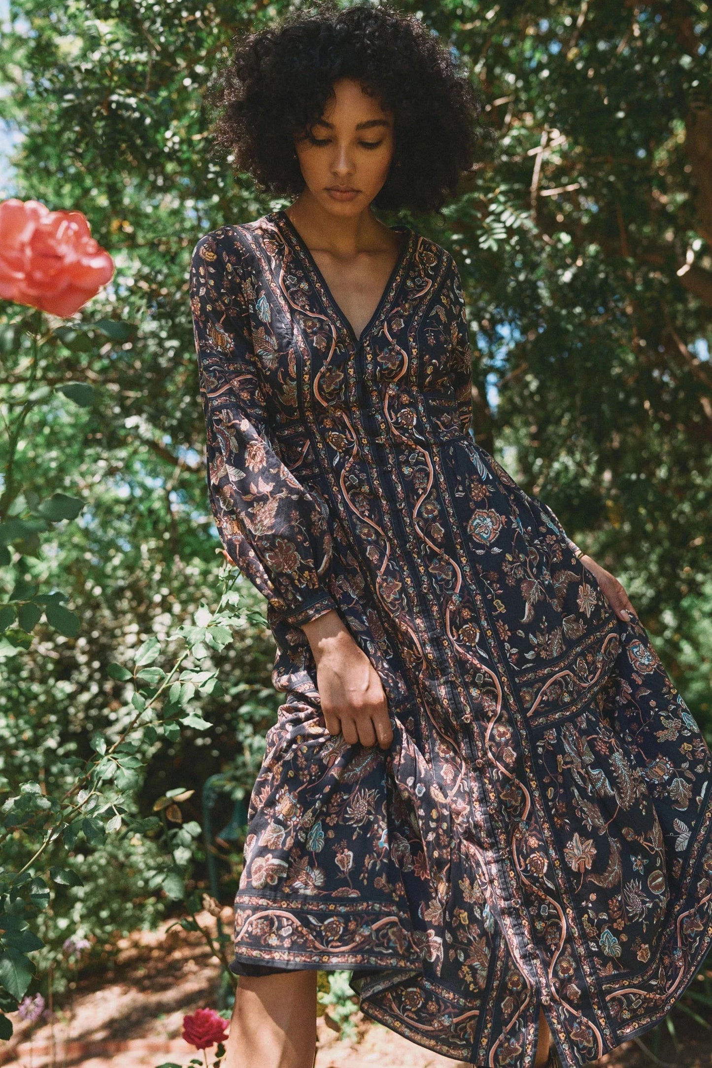 A woman wearing a long-sleeve bohemian-style maxi dress with a dark navy blue base and intricate paisley patterns in beige, brown, and muted green, standing outdoors in a garden with lush green foliage and a red rose in the background.