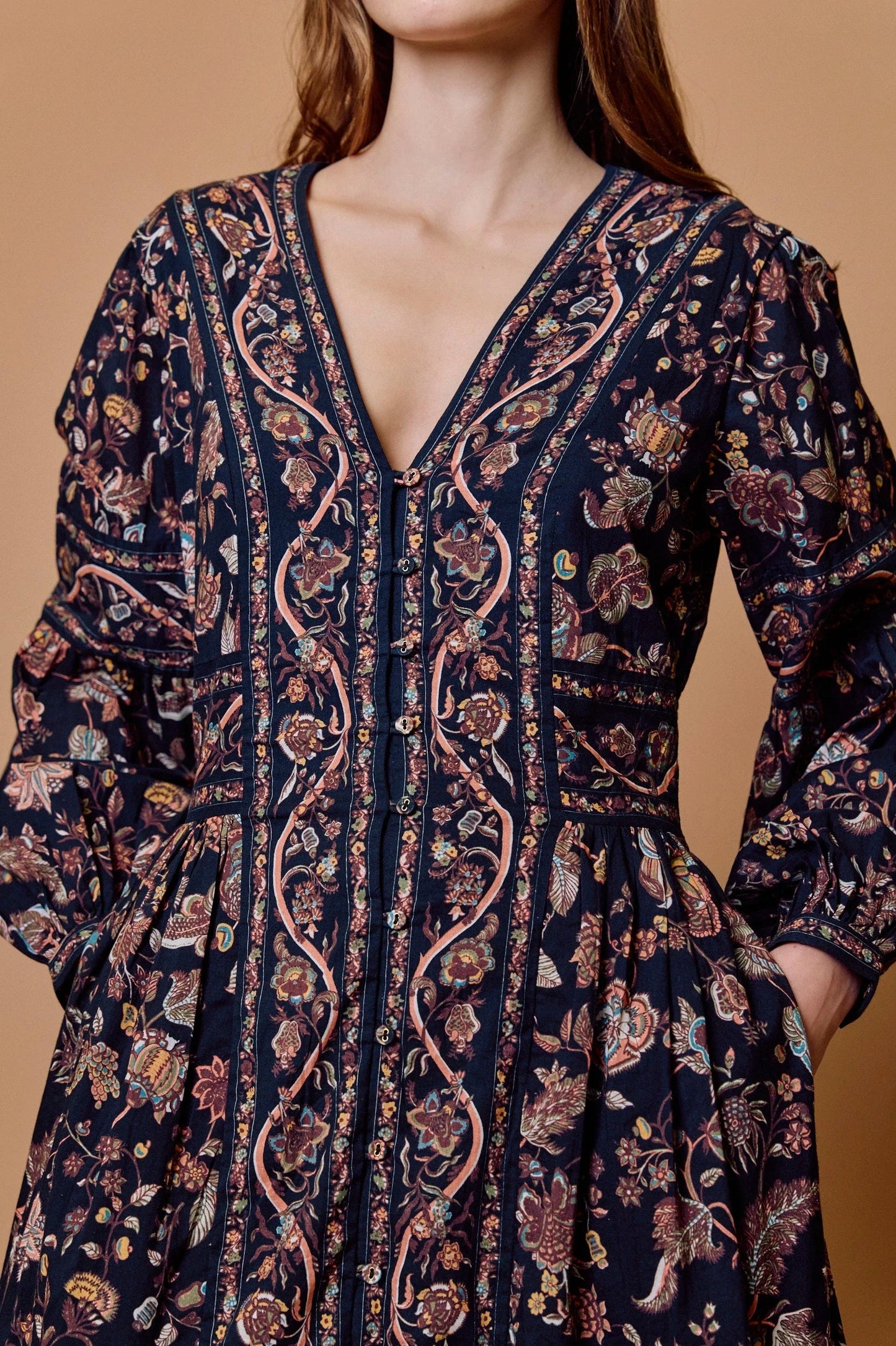 A close-up of a woman wearing a dark navy blue long-sleeve dress with a detailed bohemian floral and paisley print in pink, beige, and brown, set against a solid warm tan background.