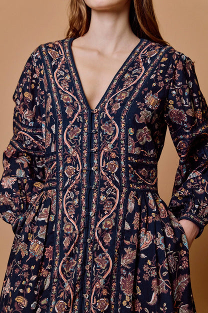 A close-up of a woman wearing a dark navy blue long-sleeve dress with a detailed bohemian floral and paisley print in pink, beige, and brown, set against a solid warm tan background.