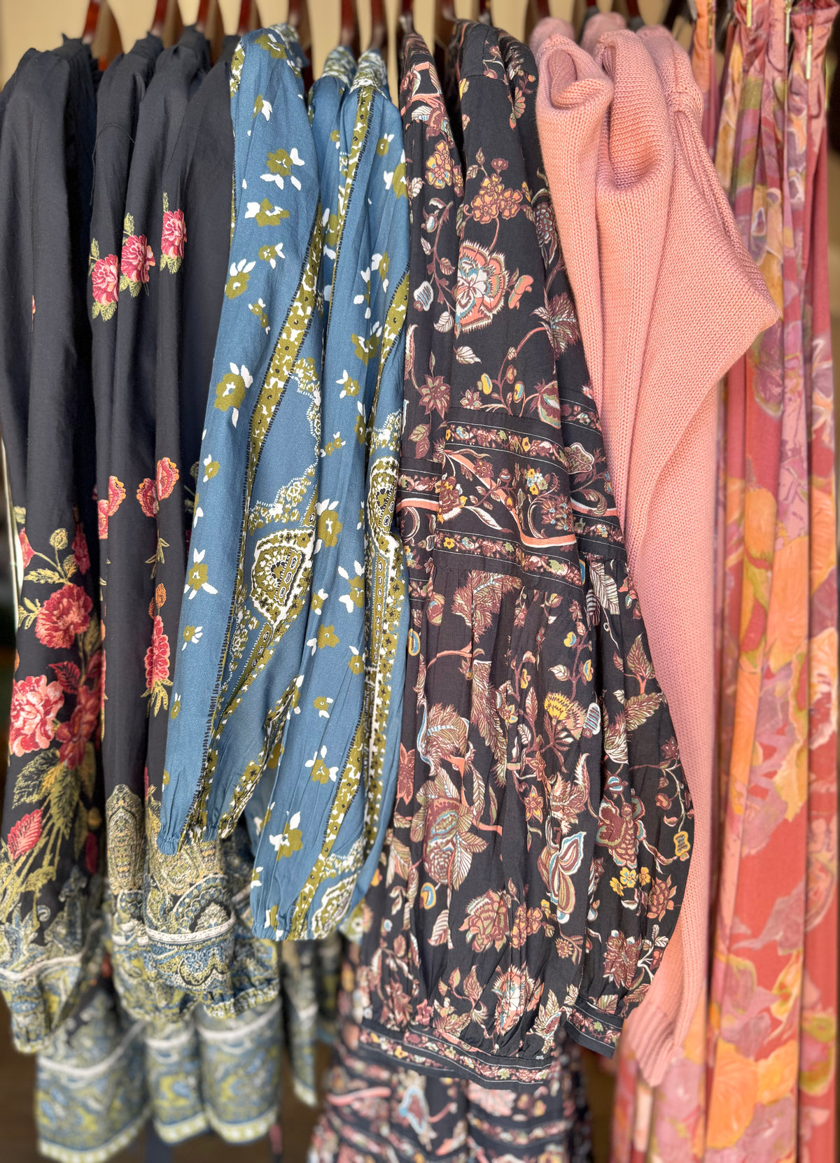 A close-up row of hanging garments in different prints and colors, including dark floral, blue paisley with gold trim, black floral, pink knit, and orange floral patterns.