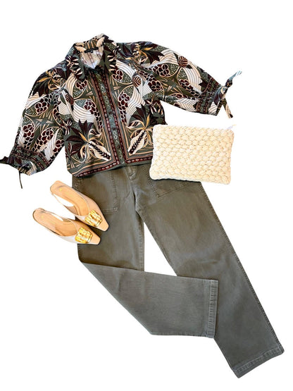 A styled flat-lay outfit featuring a black batik patterned shirt jacket, olive green pants, a cream woven clutch, and beige pointed-toe flats with gold accents, arranged on a solid white background.