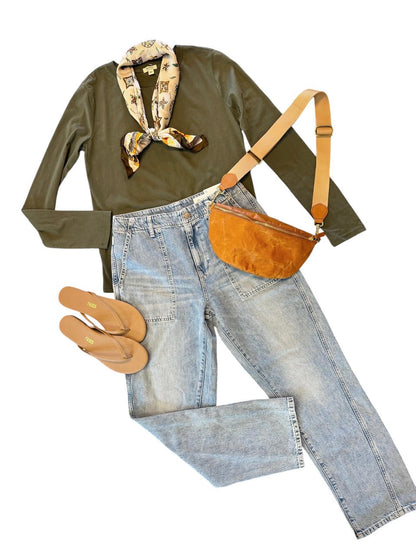 A styled flat-lay outfit on a white background featuring a dark olive green long-sleeve shirt with a patterned neck scarf, light-wash blue jeans, tan sandals, a brown suede crossbody bag, and a matching brown leather belt.